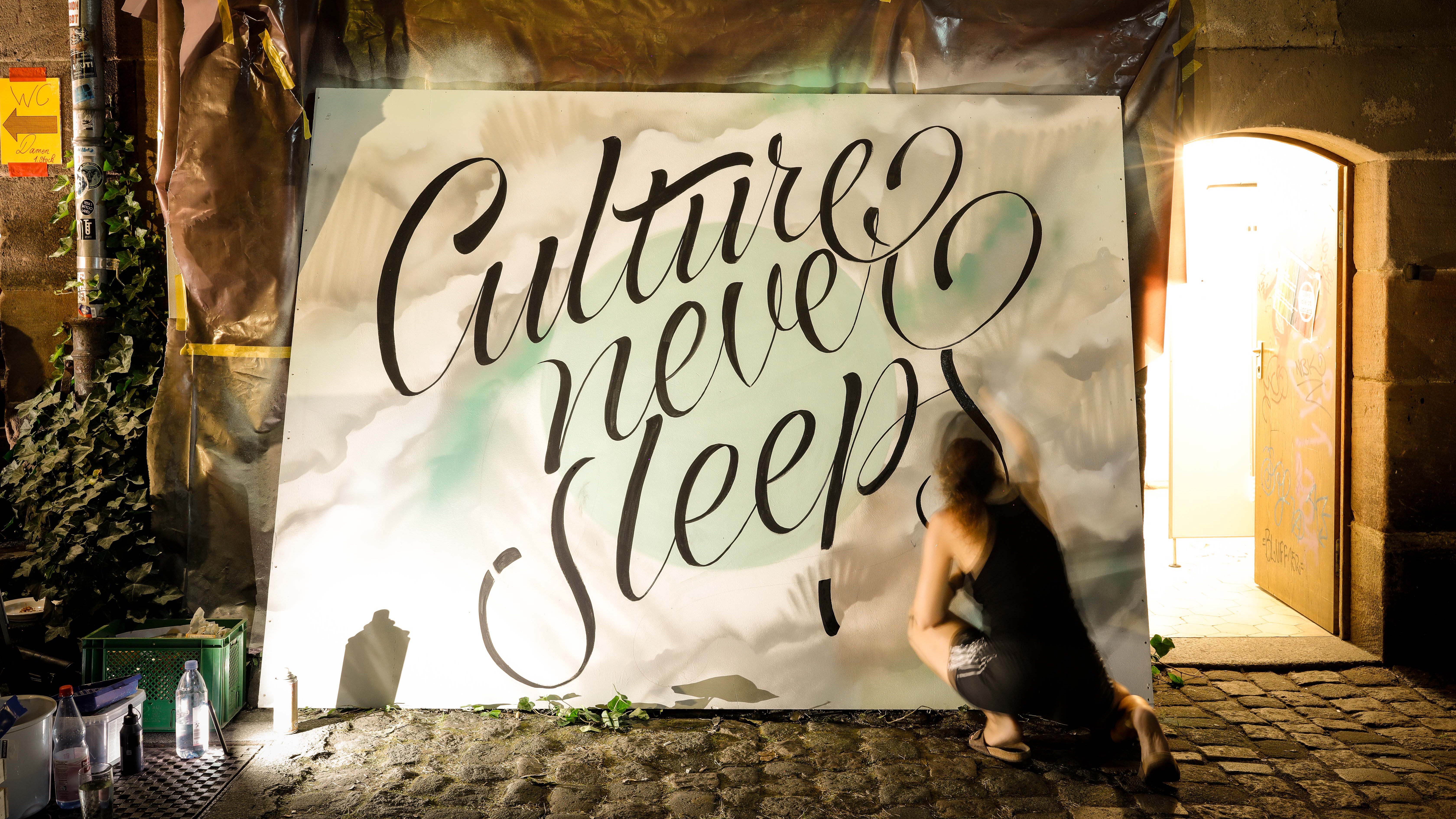 Frau kniet vor großer Leinwand und sprüht den Schriftzug ‚Culture never sleeps‘ in schwarzer Schreibschrift.