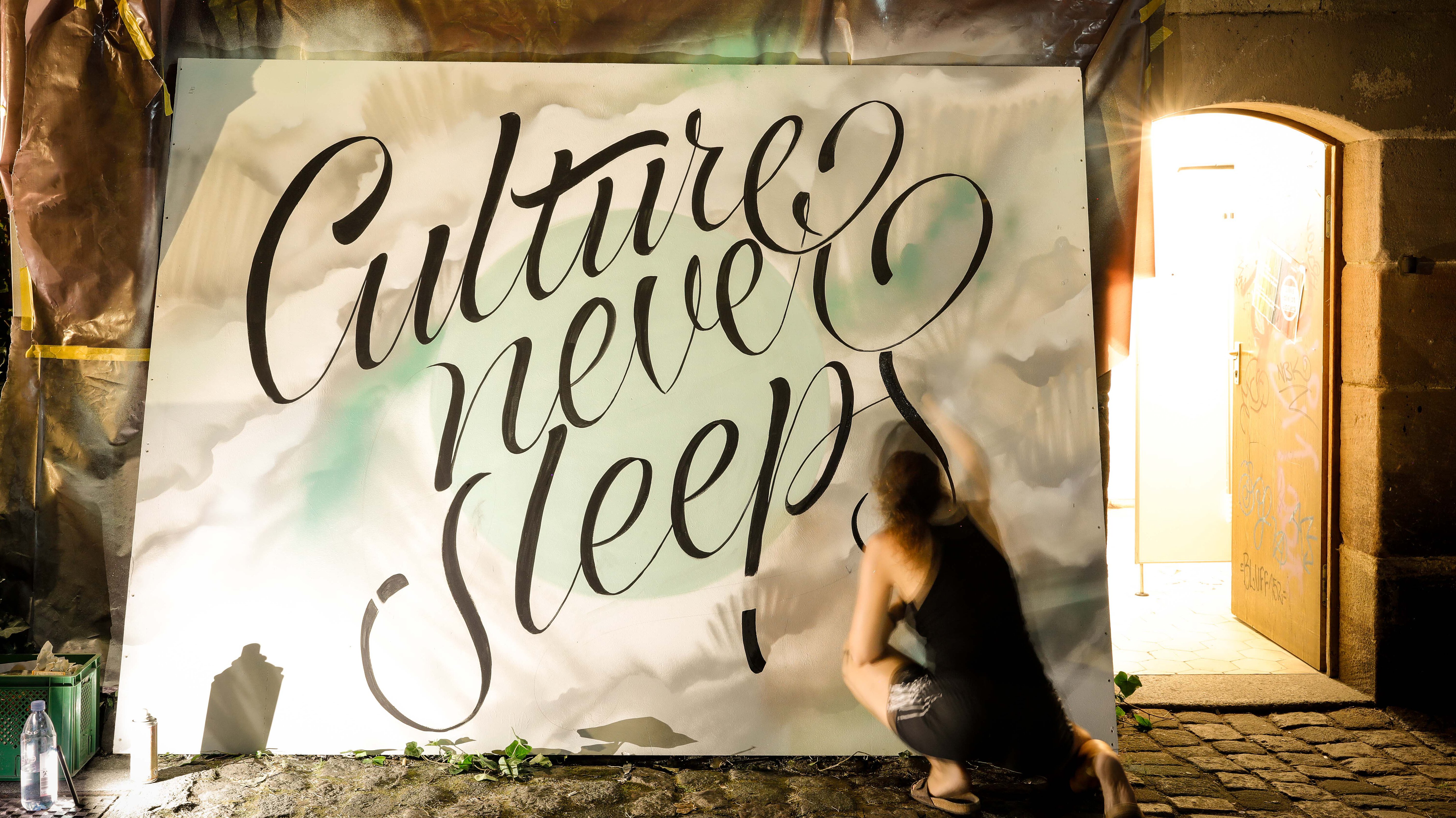 Frau kniet vor großer Leinwand und sprüht den Schriftzug ‚Culture never sleeps‘ in schwarzer Schreibschrift.
