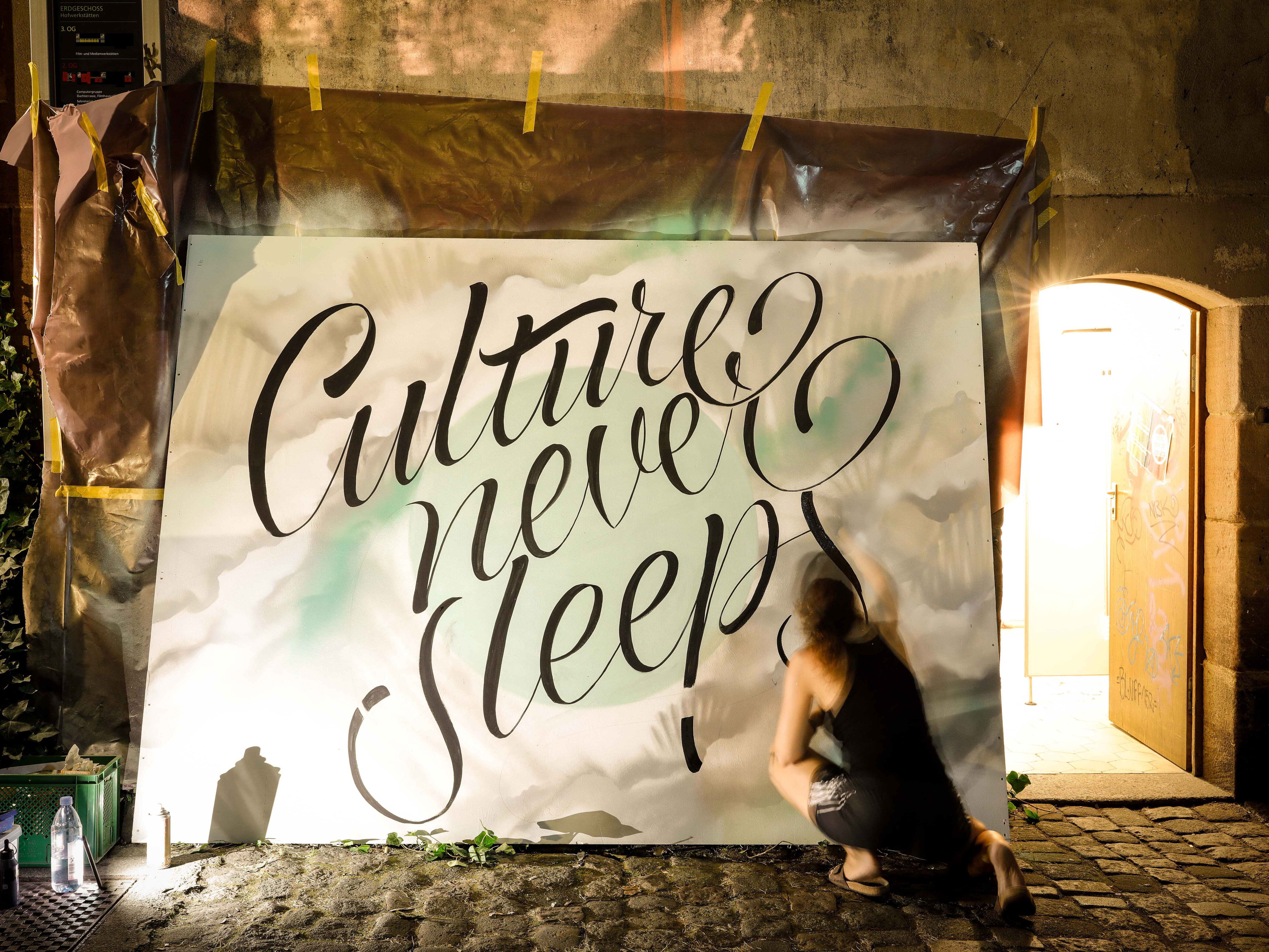 Frau kniet vor großer Leinwand und sprüht den Schriftzug ‚Culture never sleeps‘ in schwarzer Schreibschrift.