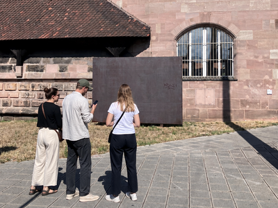 Drei Personen stehen vor einem großen, rechteckigen Kunstwerk aus dunklem Material vor einem historischen Gebäude mit roten Ziegeldächern.