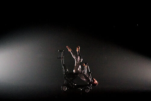 Person in enger Kleidung liegt rückwärts auf einem Rollstuhl mit hochgestreckten Beinen vor dunklem Hintergrund