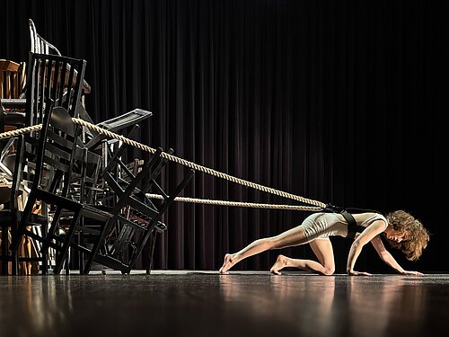 Eine Frau in Sportkleidung stützt sich auf Händen und Füßen auf einer Bühne ab, links ein Stapel aus mehreren schwarzen Stühlen, im Hintergrund ein schwarzer Vorhang.