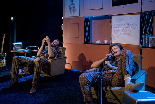 Zwei Personen in karierten Hosen sitzen auf grauen Sofas. Eine Person hält ein Smartphone, die andere hat ein Mikrofon. Im Hintergrund sind Notizen an einer Wand.