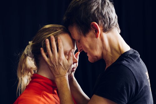 Eine Frau mit kurzen, grauen Haaren hält das Gesicht einer blonden Frau mit Zopf in beiden Händen, sie berühren einander an der Stirn