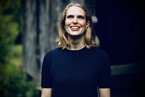 Person mit schulterlangen blonden Haaren, trägt ein schwarzes T-Shirt, steht vor unscharfem Hintergrund mit Baum und dunkler Struktur.