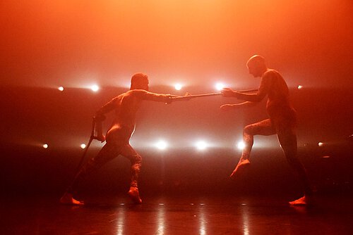 Zwei Personen stehen sich auf einer Bühne gegenüber und berühren sich mit langen Stäben, umgeben von starkem Gegenlicht und Nebel.