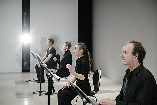 Vier Personen in schwarz gekleidet in einer Reihe auf Stühlen sitzend