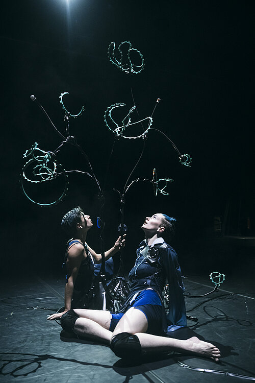 Zwi Tänzerinnen sitzen am Fuße eines Gestells mit Magnetspulen