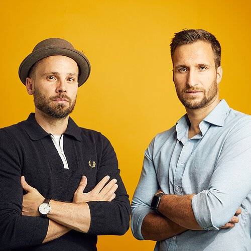 Zwei Männer mit verschränkten Armen vor orangefarbenem Hintergrund, einer trägt schwarzen Pullover und Hut, der andere hellblaues Hemd.