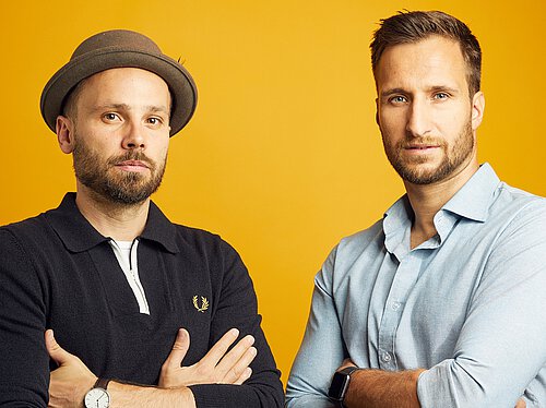 Zwei Männer mit verschränkten Armen vor gelbem Hintergrund, einer trägt einen Hut und eine schwarze Strickjacke, der andere ein hellblaues Hemd und eine Smartwatch.