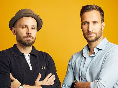 Zwei Männer mit verschränkten Armen vor orangefarbenem Hintergrund, einer trägt schwarzen Pullover und Hut, der andere hellblaues Hemd und Armbanduhr.