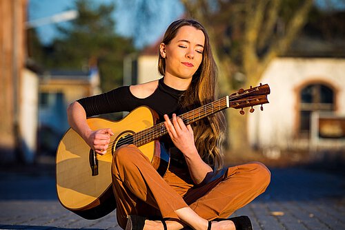 Person mit langen Haaren sitzt im Schneidersitz auf dem Boden und spielt akustische Gitarre