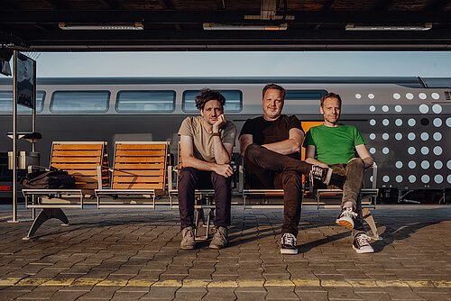 Drei Männer sitzen an einem Bahnsteig auf einer Bank, während hinter ihnen ein Zug vorbei fährt. 