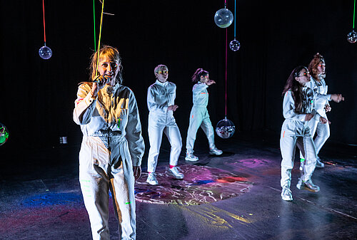 fünf Frauen in weißen Overalls mit Mikrofonen auf der Bühne