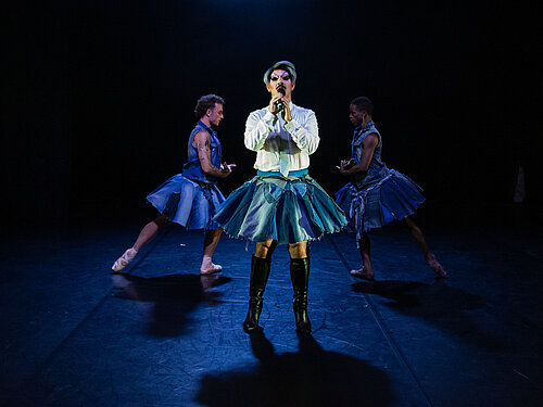 Drei Personen in schwarzen Tüllröcken und schwarzen Schuhen stehen eng beieinander auf einer Bühne mit blauem Licht und Nebel, davor eine Discokugel.