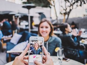 Junge Frau sitzt lächelnd an einem Tisch im Café und wird von einer anderen Person mit dem Smartphone fotografiert