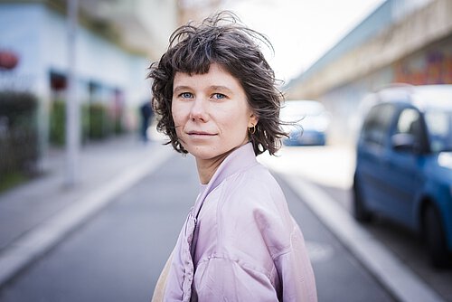 Frau mit kurzen, gewellten Haaren und hellviolettem Mantel steht auf einer Straße mit unscharfem Hintergrund und blickt in die Kamera