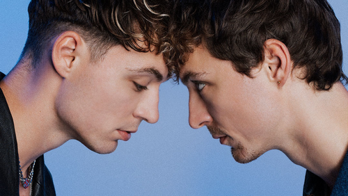 Zwei junge Männer stehen sich gegenüber, beide tragen Halsketten und haben kurze Haare