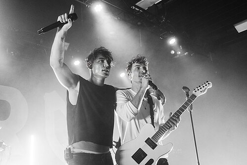Zwei Musiker auf einer Bühne, einer hält ein Mikrofon und hebt den Arm, der andere spielt eine E-Gitarre und singt ins Mikrofon