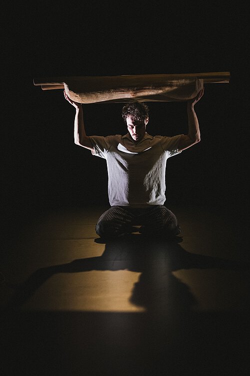 Ein Mann sitzt im Gegenlicht, er hebt eine Holzmolle mit rechtwinkligen Armen über seinen Kopf