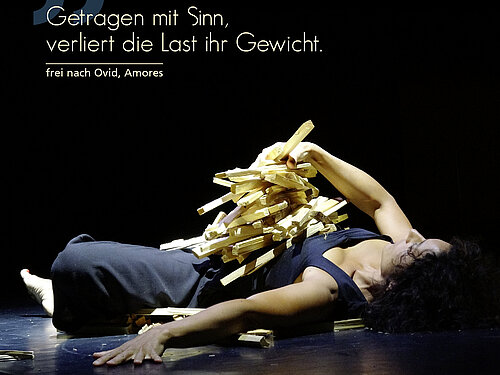 Person liegt auf dem Rücken auf einer Bühne, mit ausgestrecktem Arm und Holzklötzen auf dem Oberkörper.