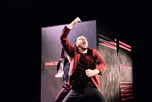 Ein Tänzer in einem roten Anzug mit schwarzem Hemd zeigt eine kraftvolle Pose mit erhobenem Arm. Im Hintergrund ist ein schwarzes Bühnenobjekt mit roten Streifen sichtbar.
