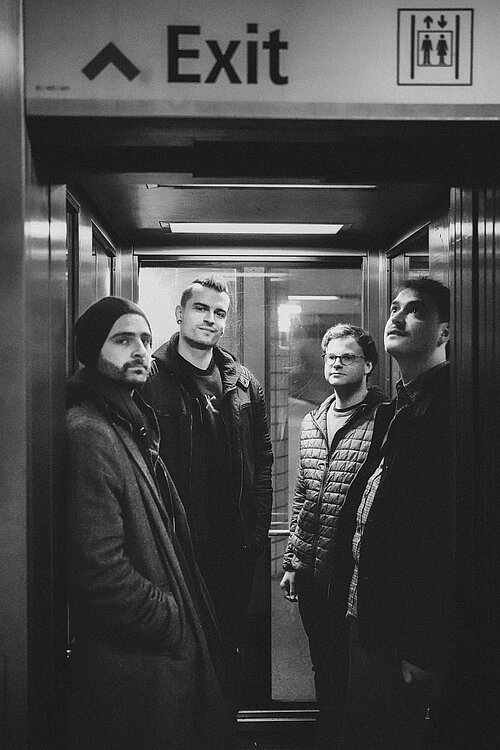 In dem schwarz-weiß Bild stehen vier Männer in einem Aufzug. 