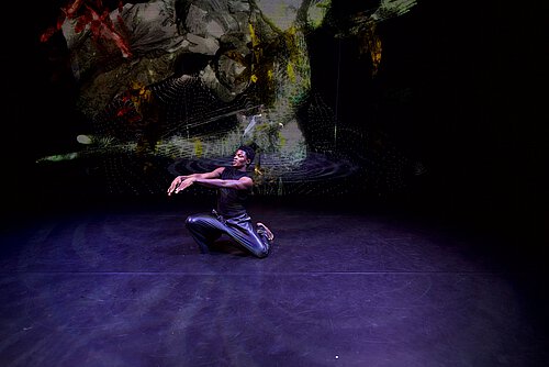 Mann mit schwarzem Overall und Dreadlocks tanzt auf schwarzem Bühnenboden, an der hinteren Bühnenwand grüne Projektionen