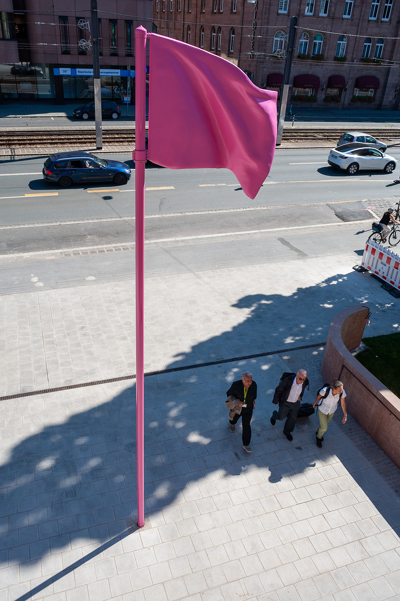 Luftaufnahme der pinkfarbenen Skulptur einer wehenden Flagge von Heidi Sill vor dem Eingang des Künstlerhauses mit drei Personen auf dem Gehweg darunter