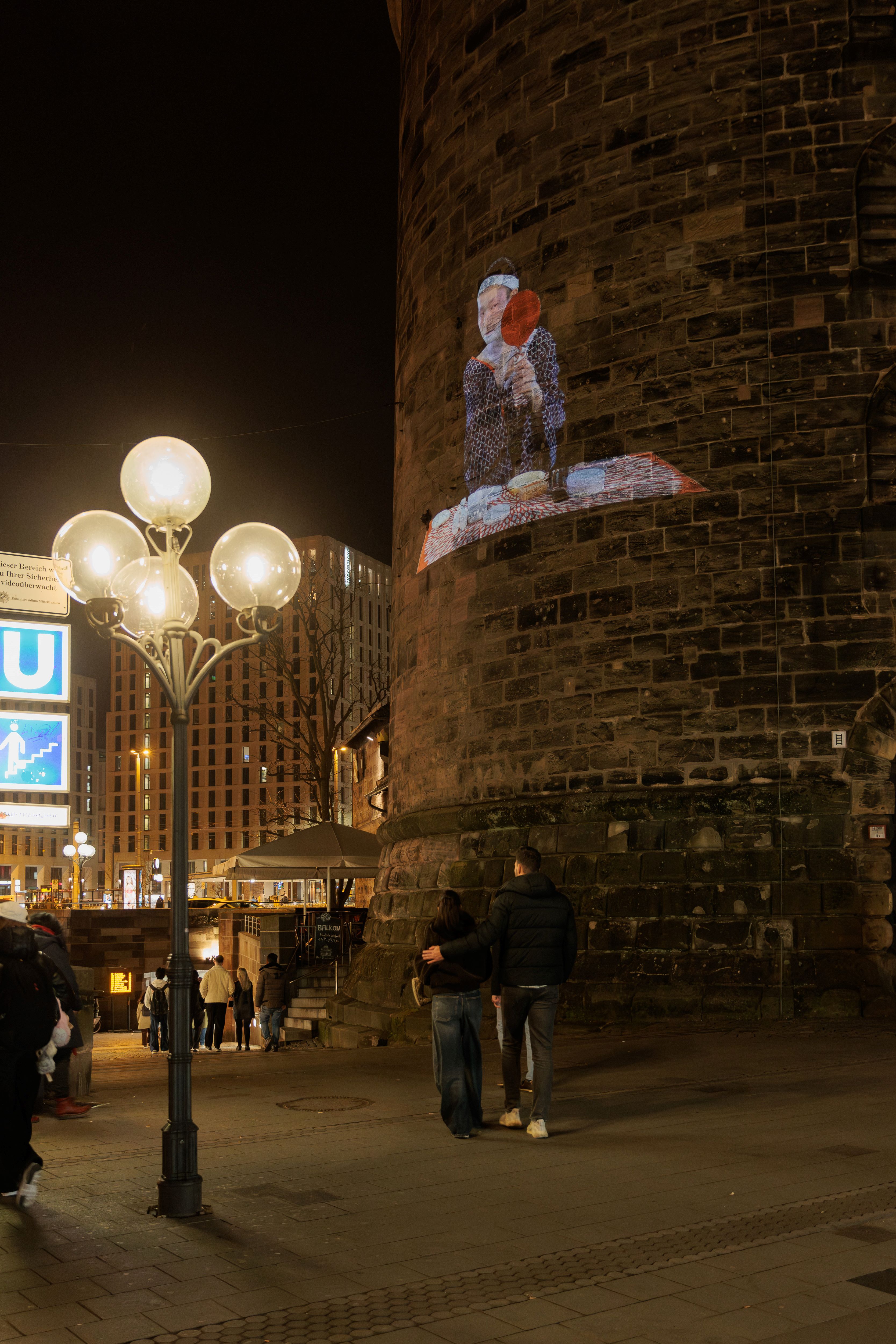 Personengruppe vor einem runden, mit Ziegeln verkleideten Turm mit einem projizierten Kunstwerk einer sitzenden Person mit rotem Fächer