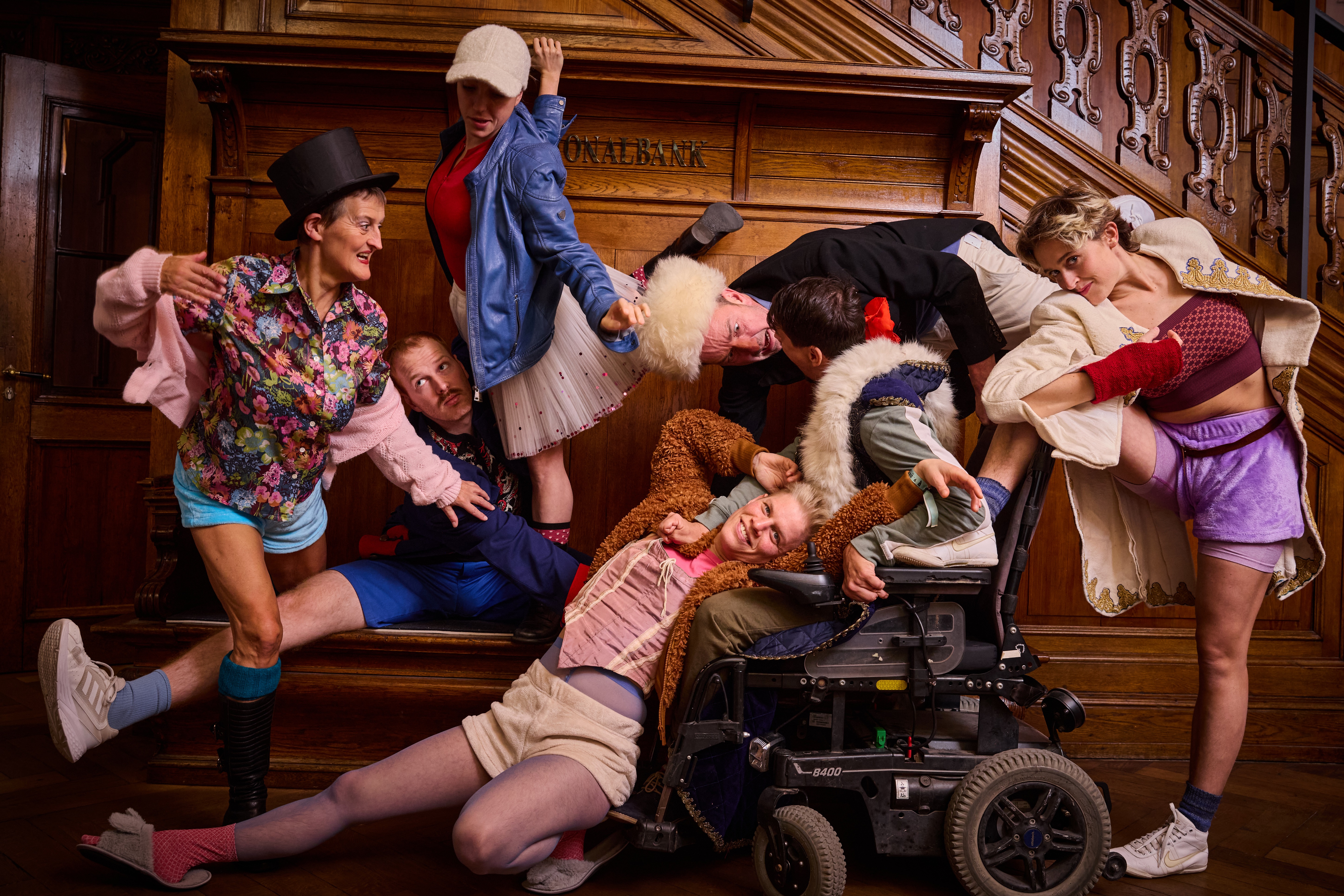 Gruppe von sieben Personen in bunten, teils historischen und modernen Kostümen vor einer Holztreppe in einem prunkvollen Innenraum.