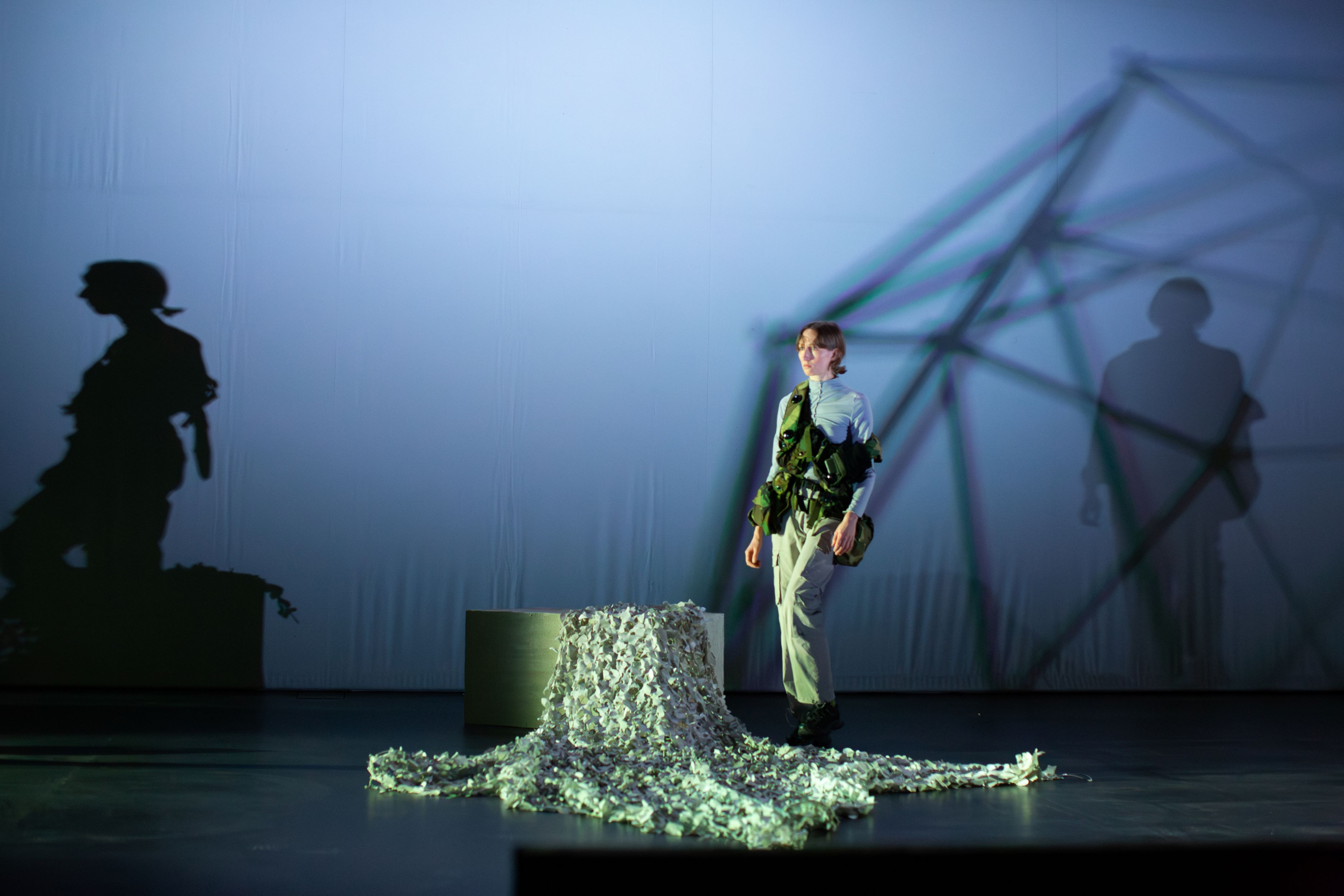 Person mit einer militärischen Weste, die über die Bühne geht.   Im Hintergrund eine weiße Leinwand, auf die Schatten der Person und einer geometrischen Form geworfen werden. Die Beleuchtung ist hell und diffus, mit einem bläulichen Farbton. Die Person schaut fokussiert in eine Richtung. Die Atmosphäre ist düster und ernst.