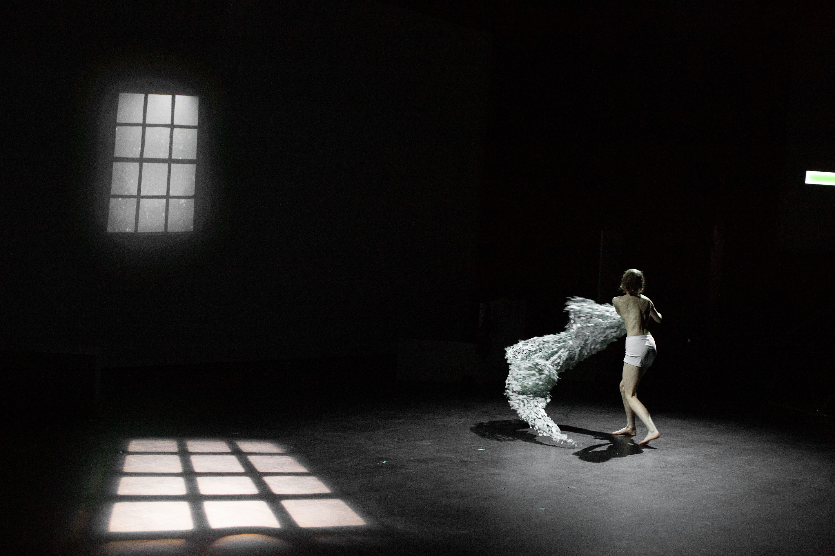 Person in Unterwäsche bewegt sich auf einer dunklen Bühne mit einem großen, weißen, abstrakten Objekt neben sich und einem Fensterlichtmuster auf dem Boden