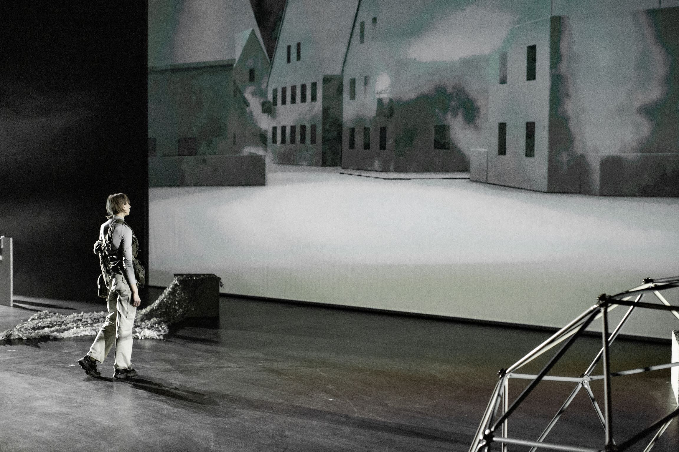 Person mit Rucksack steht auf einer Bühne vor einer großflächigen Projektion von Häusern