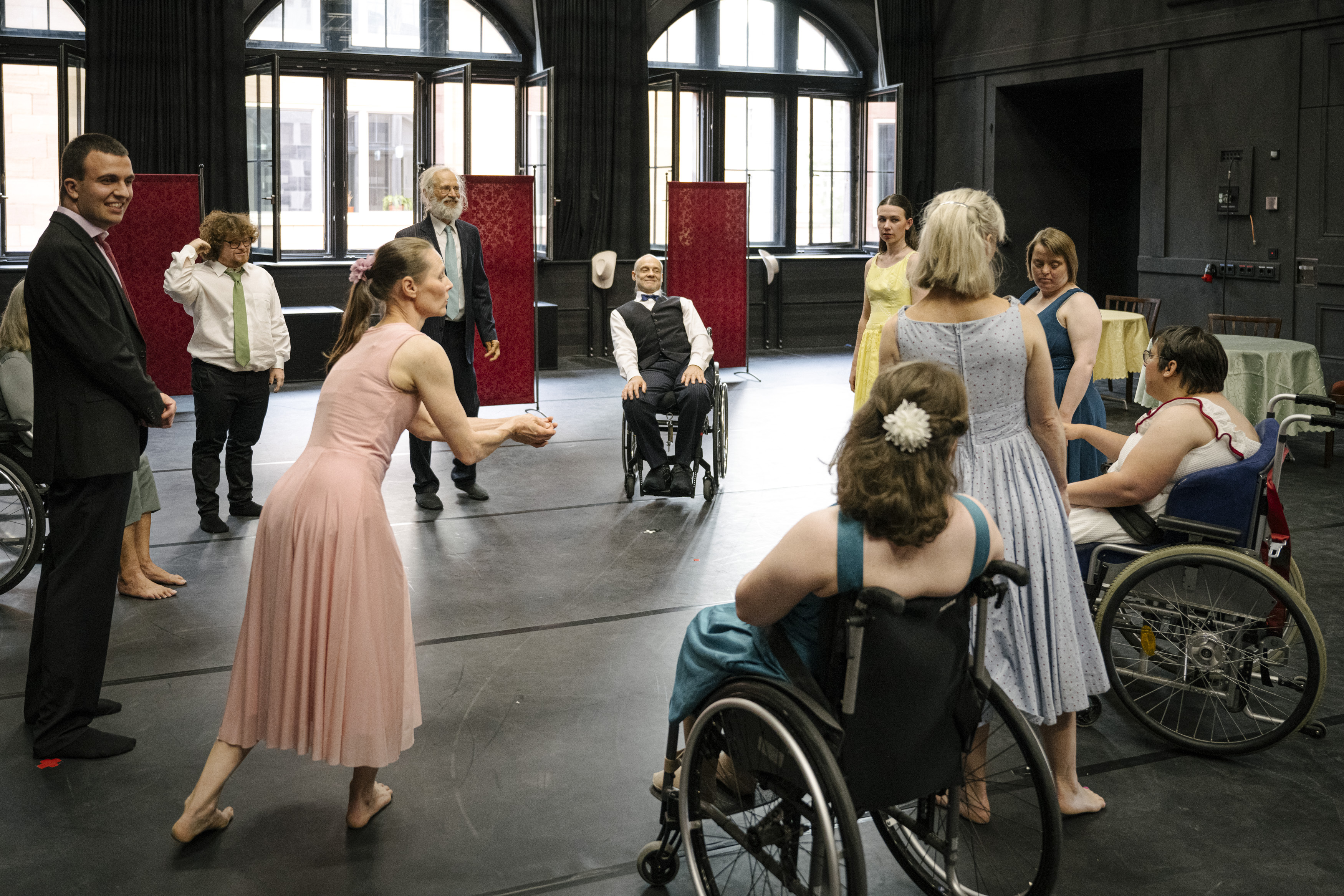 Gruppe von Menschen, darunter mehrere im Rollstuhl, in einem großen Raum mit hohen Fenstern, einige tragen festliche Kleidung und eine Frau in einem rosa Kleid macht eine einladende Geste.