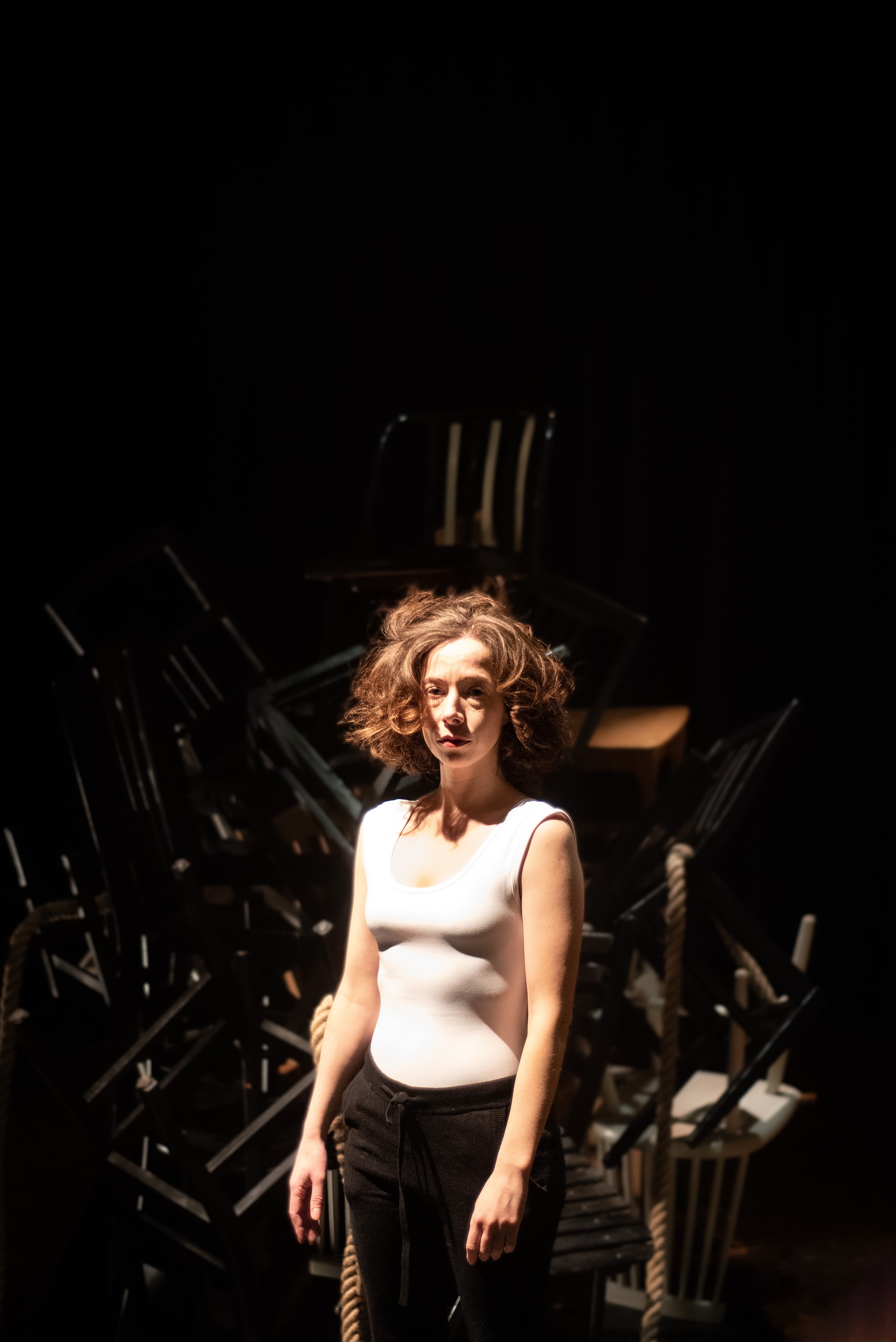 Frau mit lockigem Haar und weißem Oberteil steht vor einem dunklen Hintergrund mit einem Haufen übereinander gestapelter Stühle