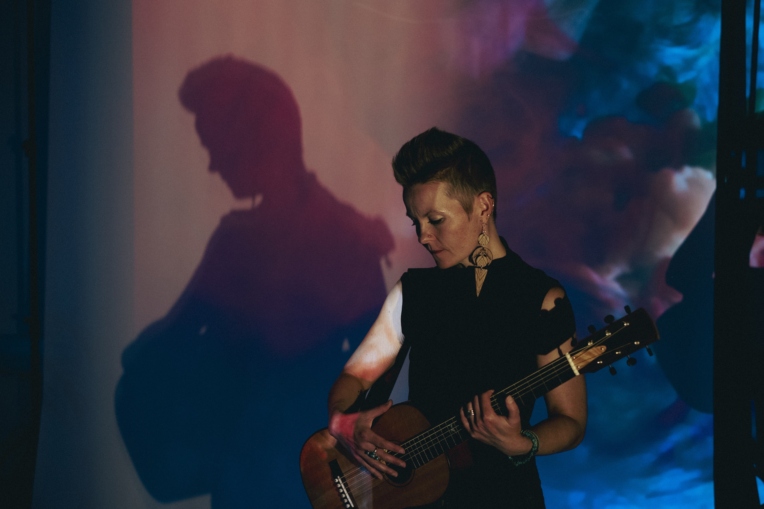 Person mit kurzem Haar spielt akustische Gitarre vor einer Wand mit Schatten und bunten Lichtprojektionen.