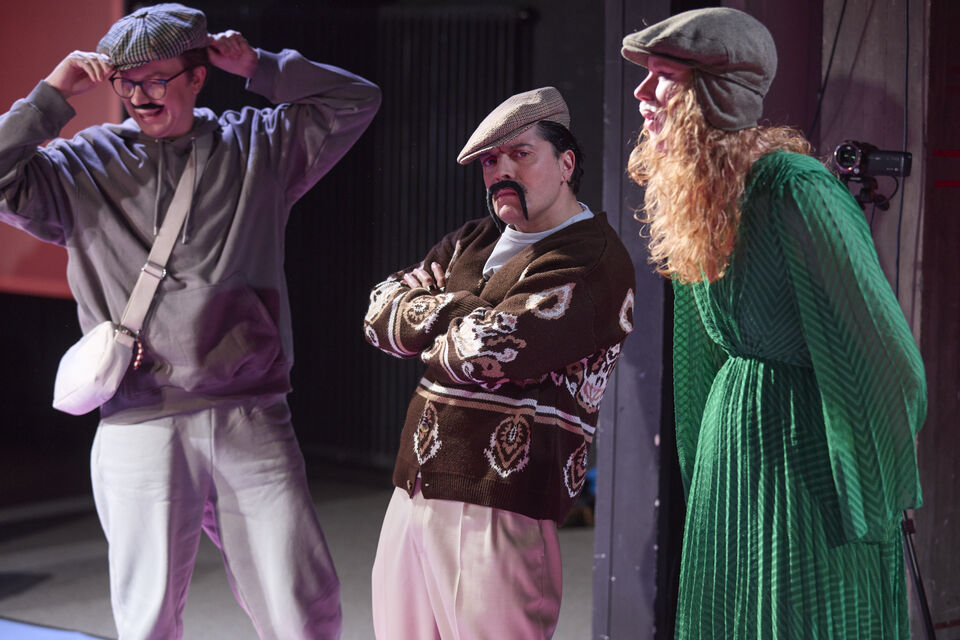 Drei Personen in Bühnenkleidung, eine mit langem lockigem Haar und Mütze, stehen in einer Theaterkulisse.