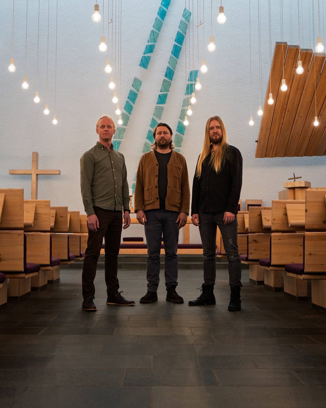 Drei Personen stehen nebeneinander in einem Raum mit hängenden Glühbirnen und blauen Wanddekorationen im Hintergrund.