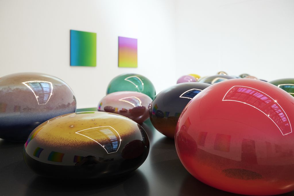 Buntfarbige runde und ellipsoide Glasobjekte auf einem Tisch, im Hintergrund Seidenmalerein in Regenbogenfarben