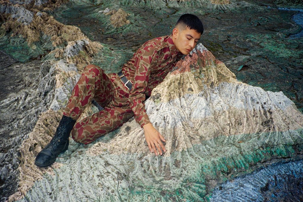 Ein Mann in Camouflagekleidung schmiegt sich an einen Fels inmitten einer steinigen Landschaft. Der Fels nimmt teilweise die Farben der Camouflage auf